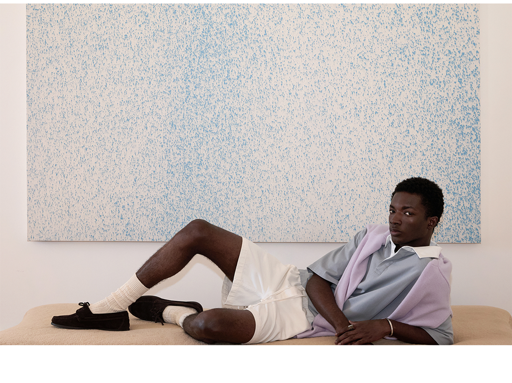 Model in blue polo and white shirts with a lilac sweater in front of a large blue and white speckle painting.