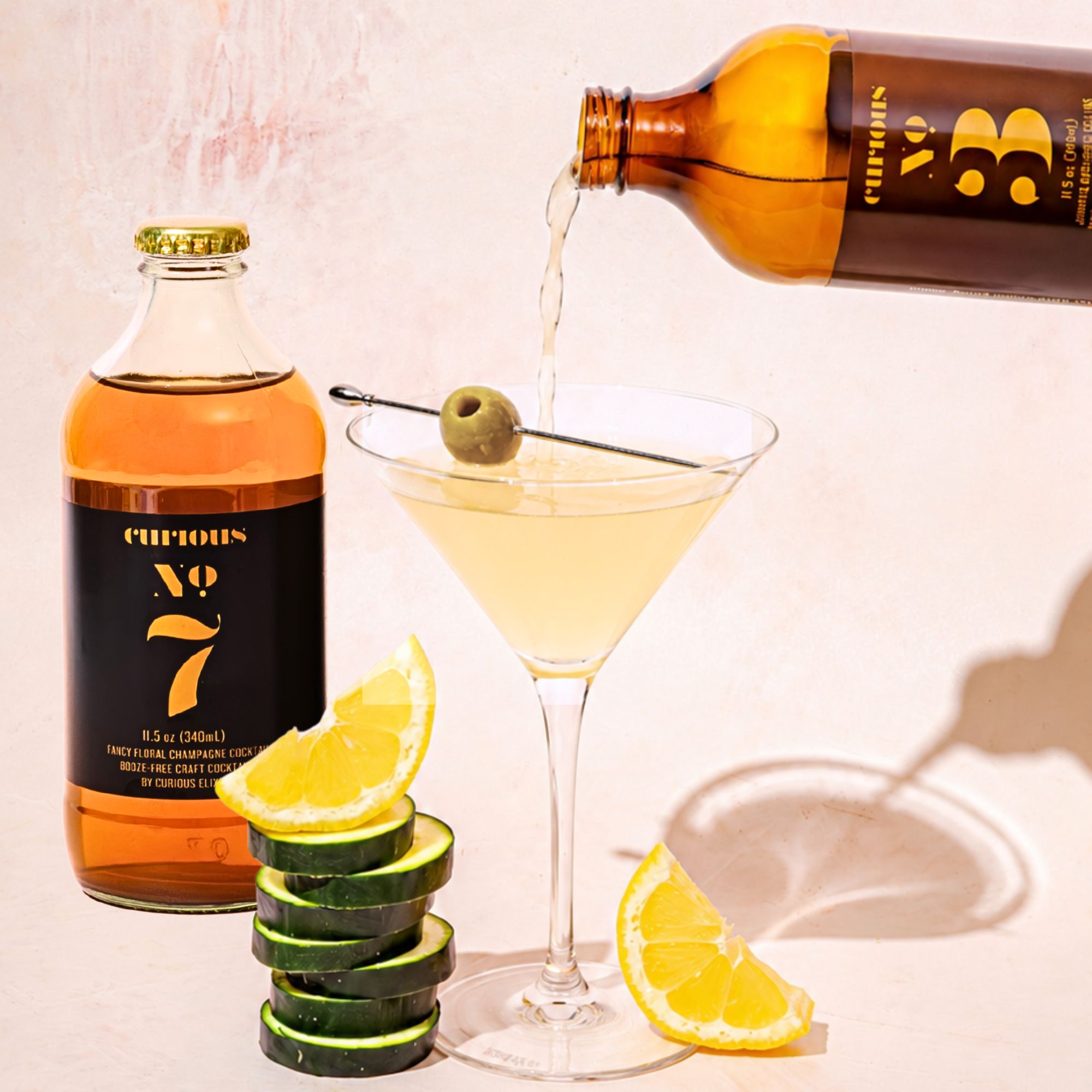 A bottle of Curious No. 3 being poured into a martini glass garnished with an olive. Slices of cucumber and lemon are stacked beside the glass. 