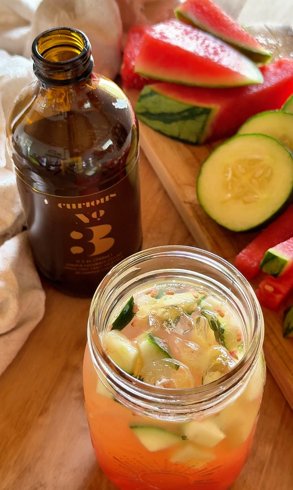 A bottle of Curious No. 3 next to a jar full of a pink drink with watermelon and cucumbers in it. Next to this is a cutting board with more watermelon and cucumbers.
