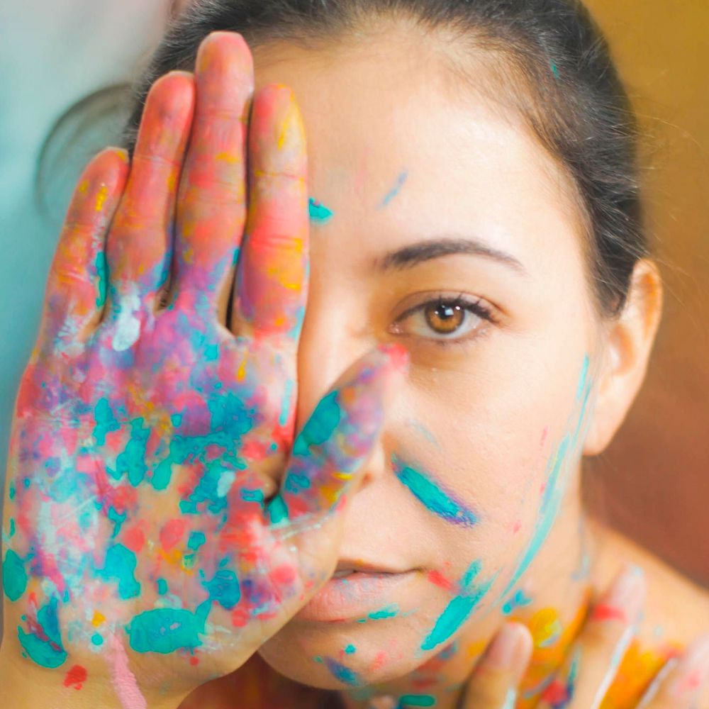 A person holds their hand up so the palm is blocking one eye. Their face and hand are covered in colorful paint. 