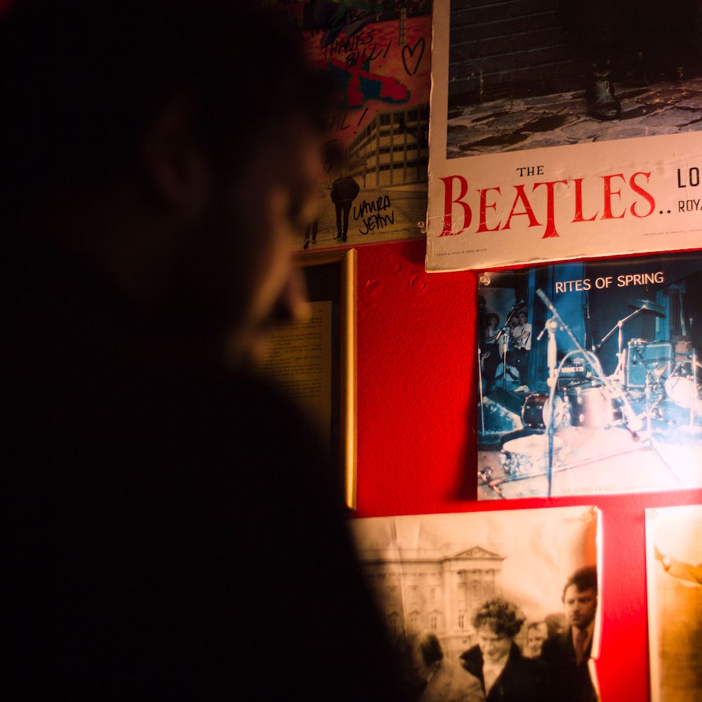 Band posters hang on a red wall in a dimly lit room, including one for The Beatles. A person, out of focus, sits in the foreground. 