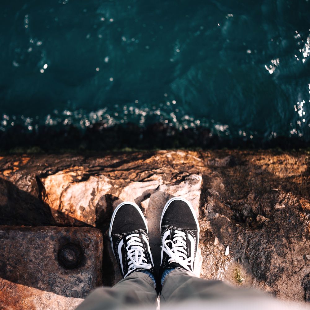 Feet wearing sneakers stand at the edge of a stone wall with a sharp drop into dark water. 